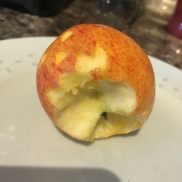 A photo of a red apple with a halloween pumpkin face carved into it. The apple sits on its side and has had a bite taken out of the bottom.